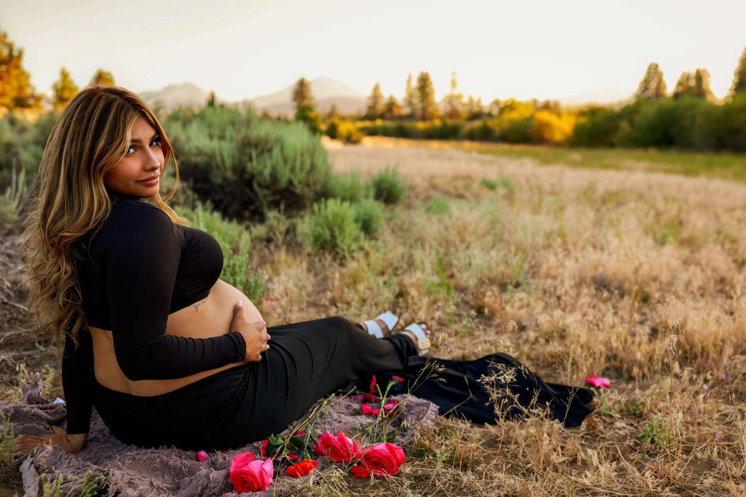 A figure in black dress sits in a golden field at sunset with red roses scattered nearby. Indian Ford Meadows, Sisters Oregon