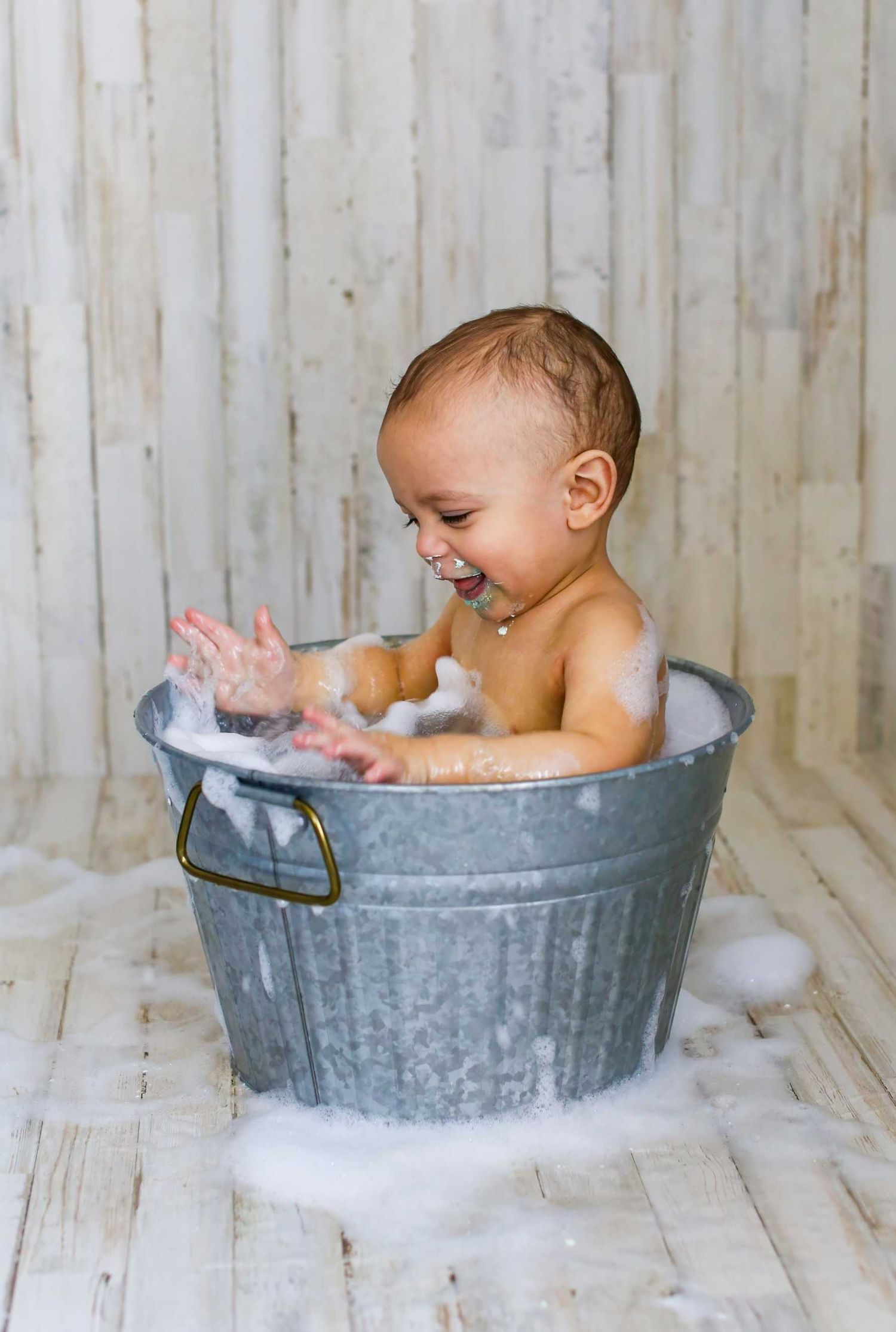 Baby splashing and playing in a vintage galvanized metal tub during bath time with bubbles and white wooden backdrop. Redmond Oregon Studio