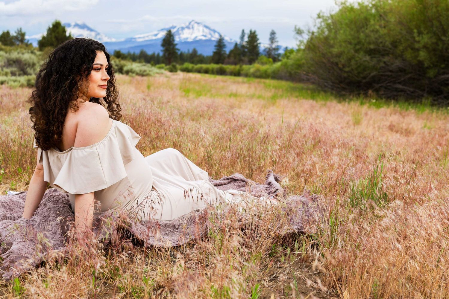 A person in a white flowy dress sits gracefully in a golden field of tall grass during sunset.