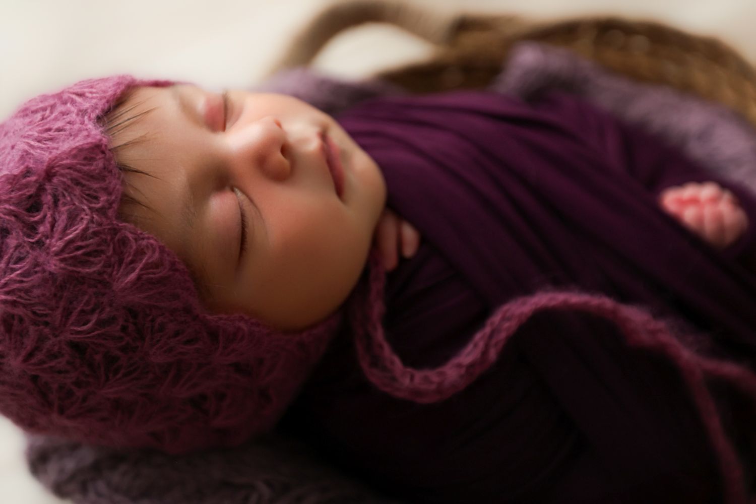 Sleeping newborn baby wrapped in purple knit blanket wearing pink crochet bonnet hat in basket.