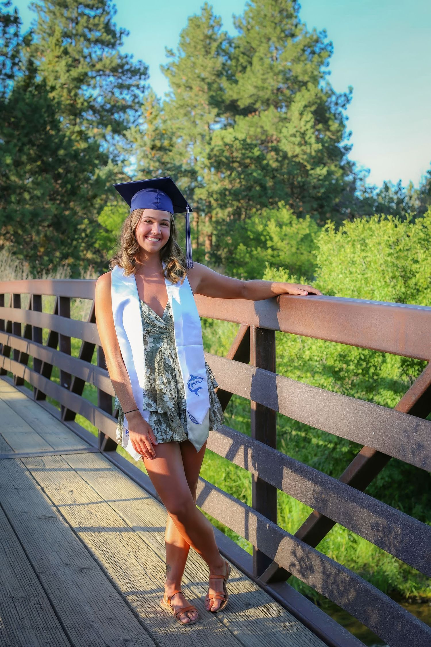 Smiling graduate in cap and white sash poses on wooden bridge surrounded by lush green trees.