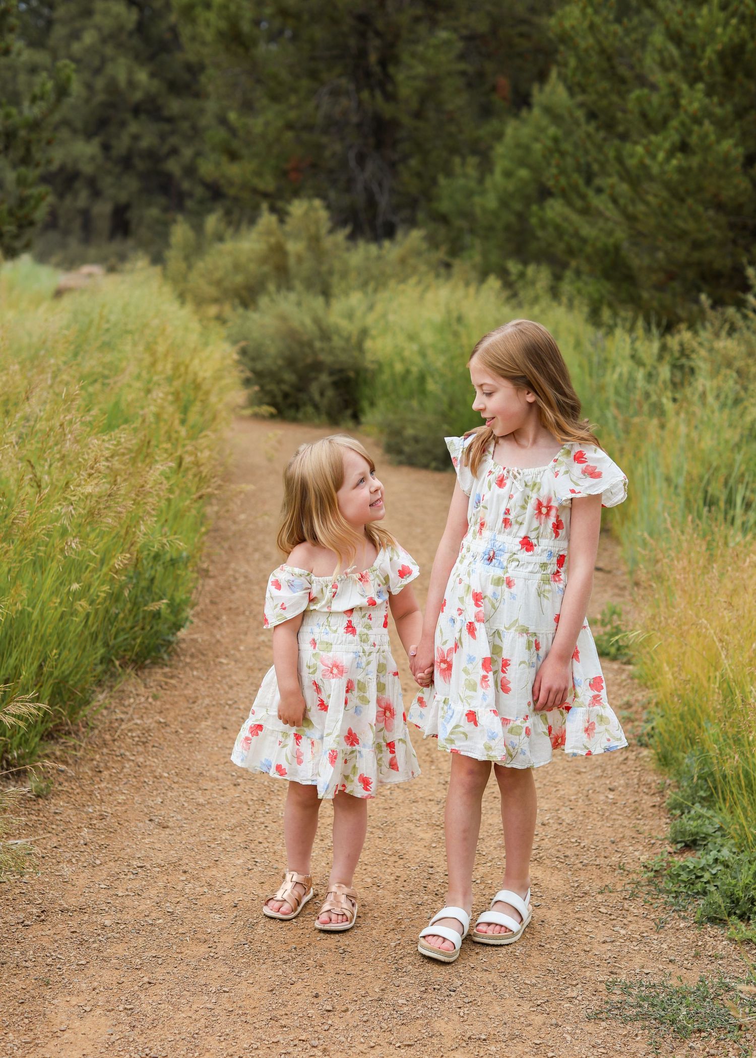 Two young girls in matching floral dresses hold hands on a dirt path surrounded by green meadow grass and trees.