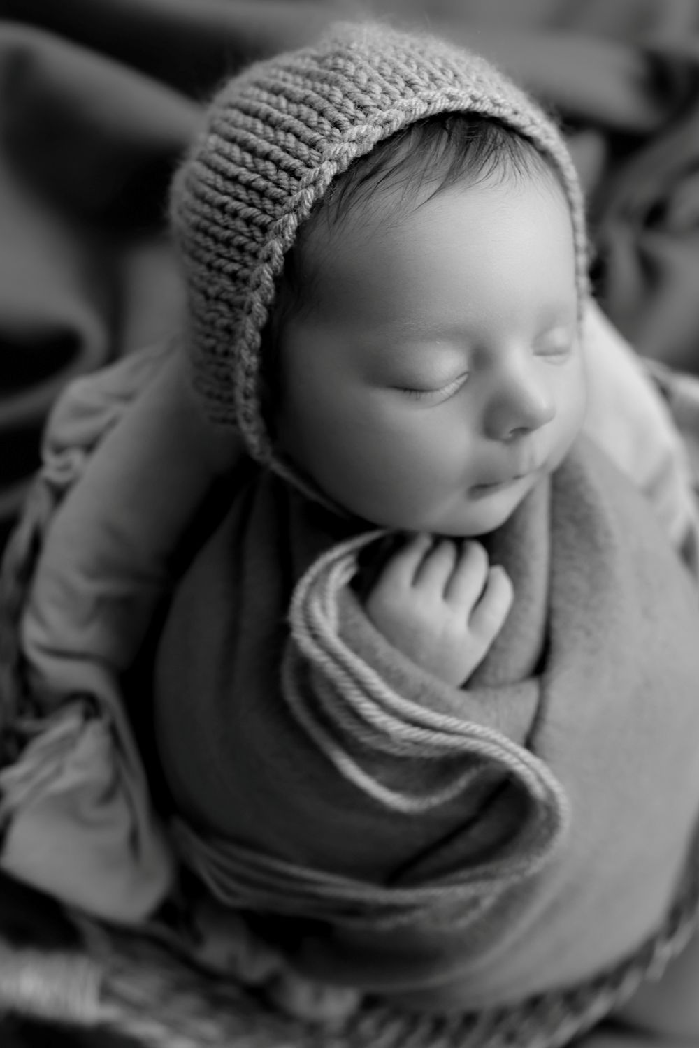 Black and white newborn photo of sleeping baby wrapped in knit blanket wearing cozy bonnet hat.