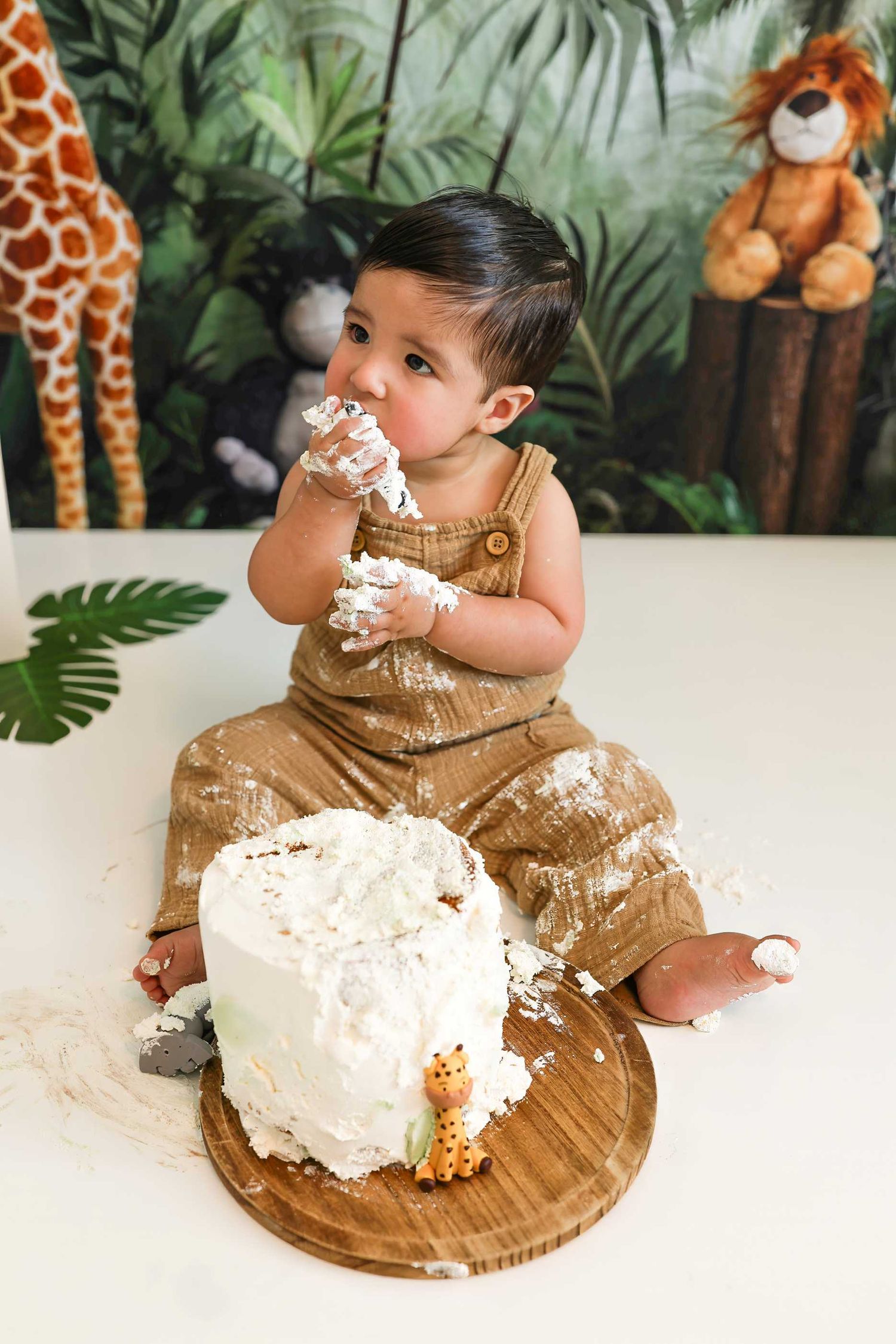Baby in brown overalls enjoys a cake smash photo session with safari-themed decorations and stuffed animals in the background.