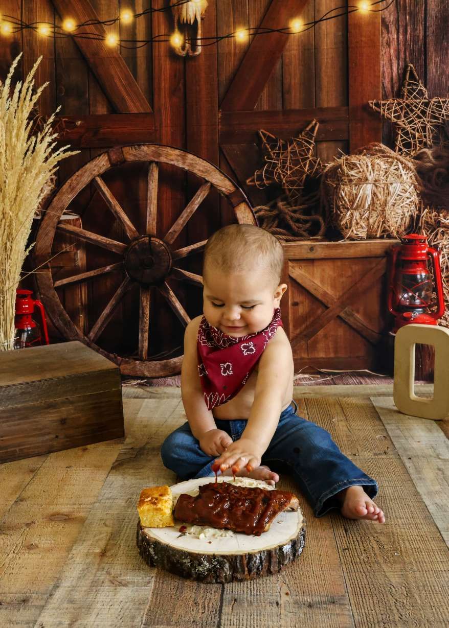 Baby boy in red bandana smashing cake on wood slice in rustic western barn birthday photo session.