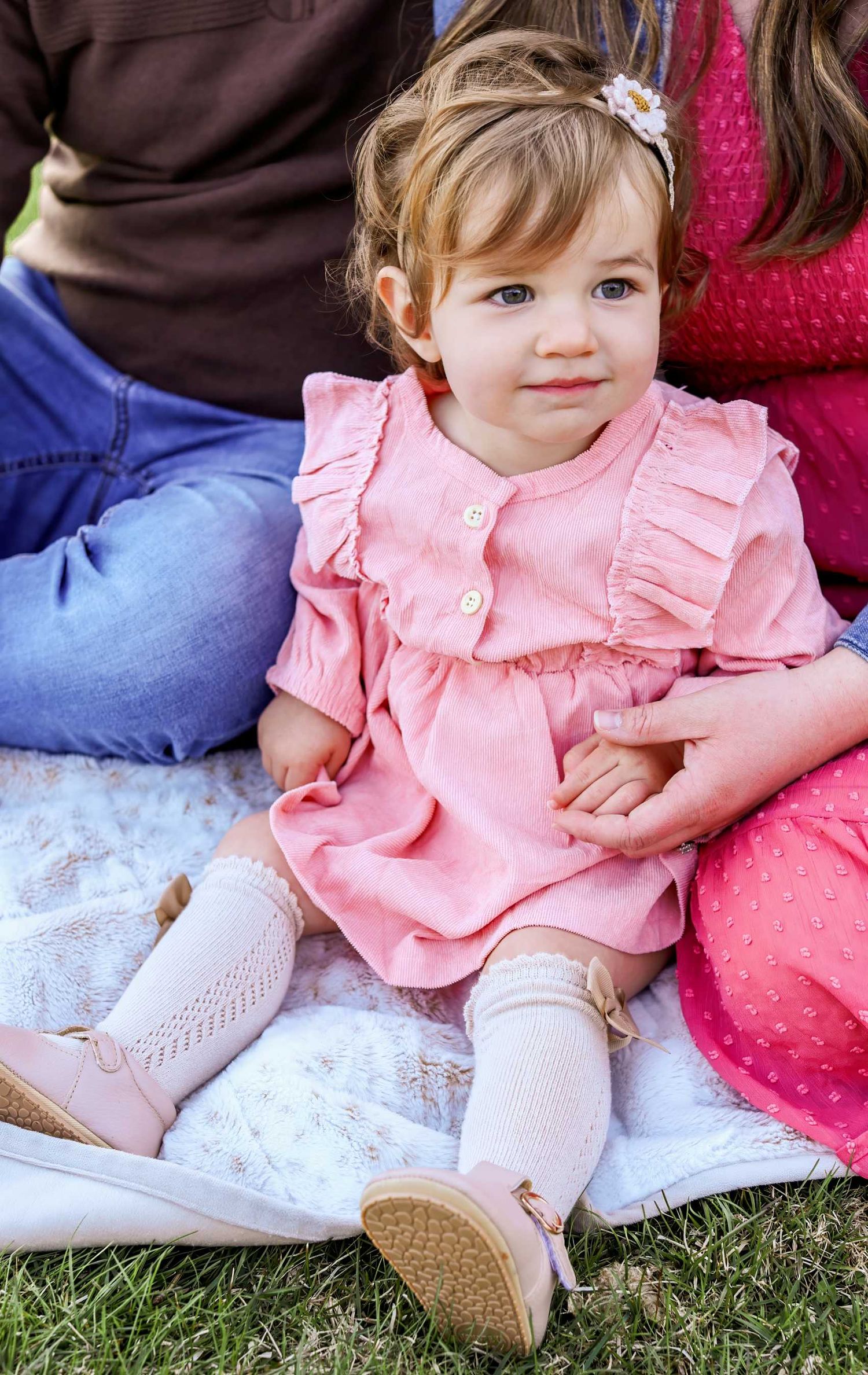 Adorable toddler girl in pink ruffle dress, white socks, and pink shoes sits on blanket outdoors with family.