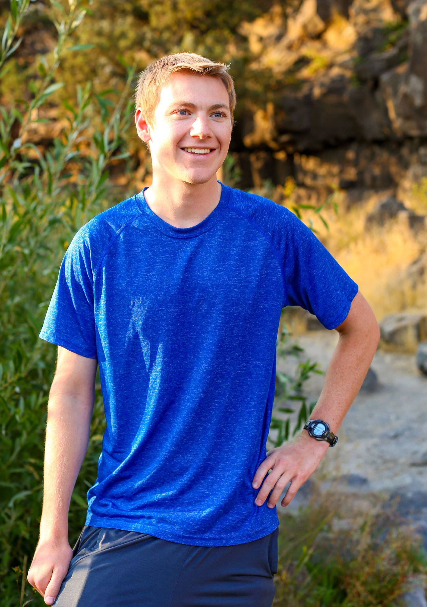 Young man smiling outdoors in blue athletic shirt and watch, posing against natural rock and greenery background.