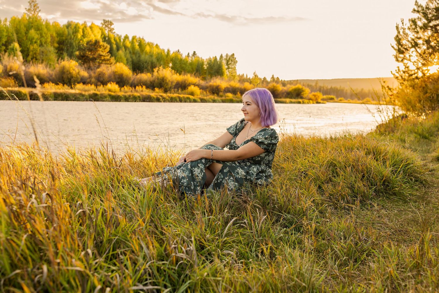 Woman with purple hair in floral dress sits in golden grass by a sunlit river at sunset.