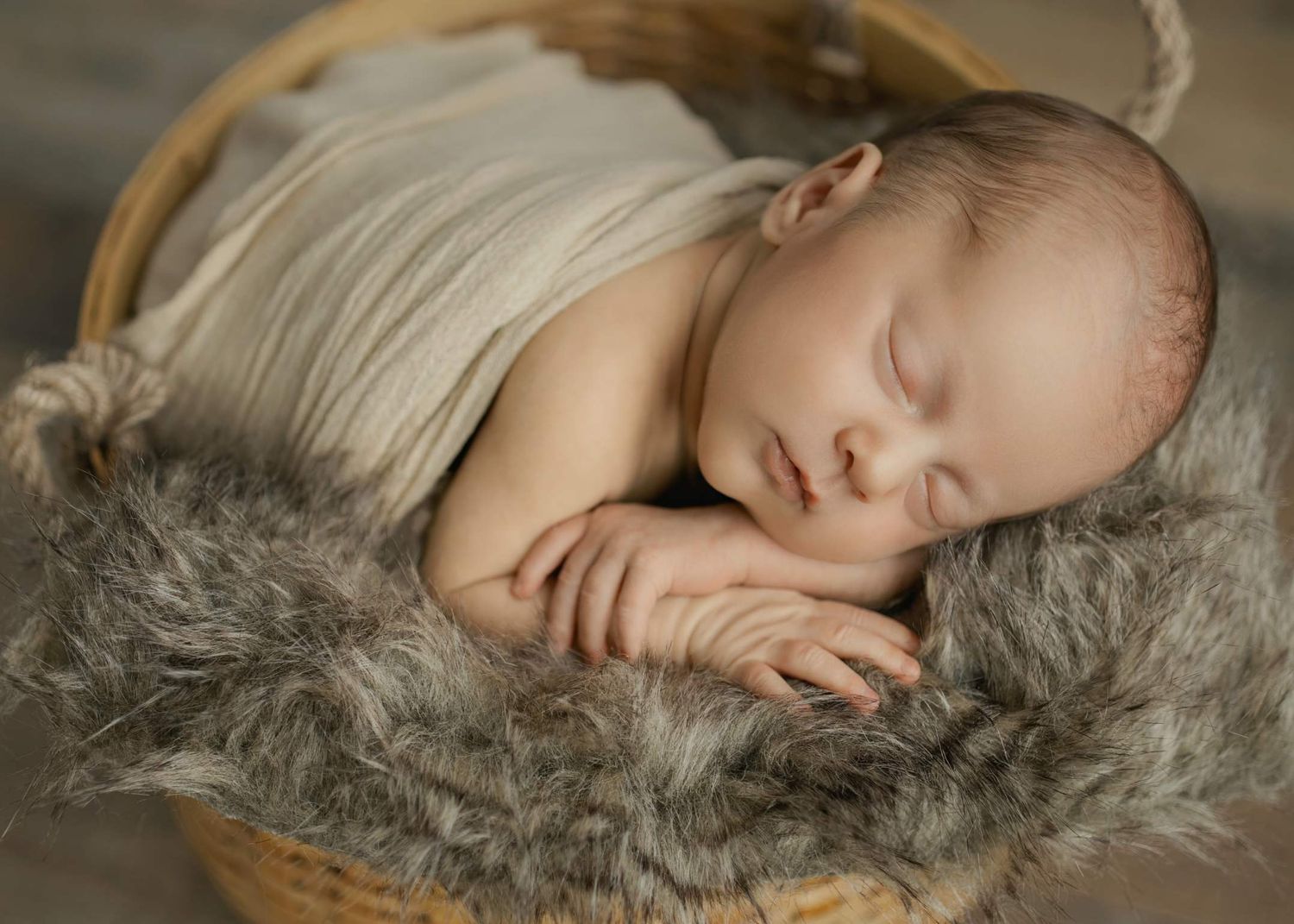 Peaceful sleeping newborn resting in wooden basket with soft gray fur lining in studio portrait setting.