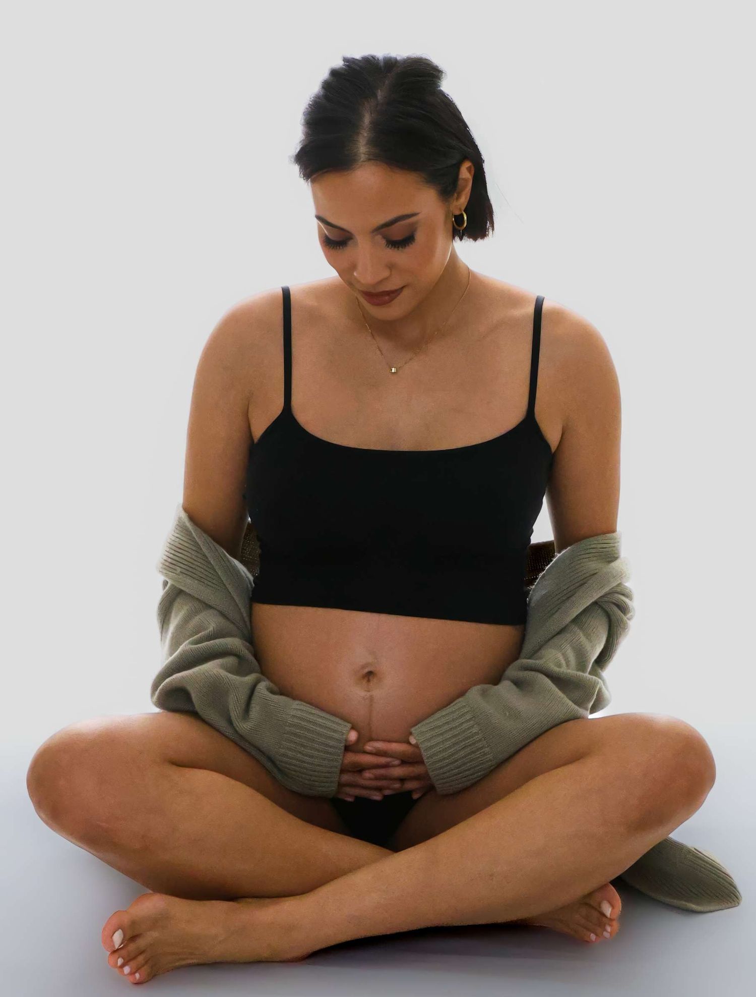 Person in black top and olive cardigan sitting cross-legged against white background.