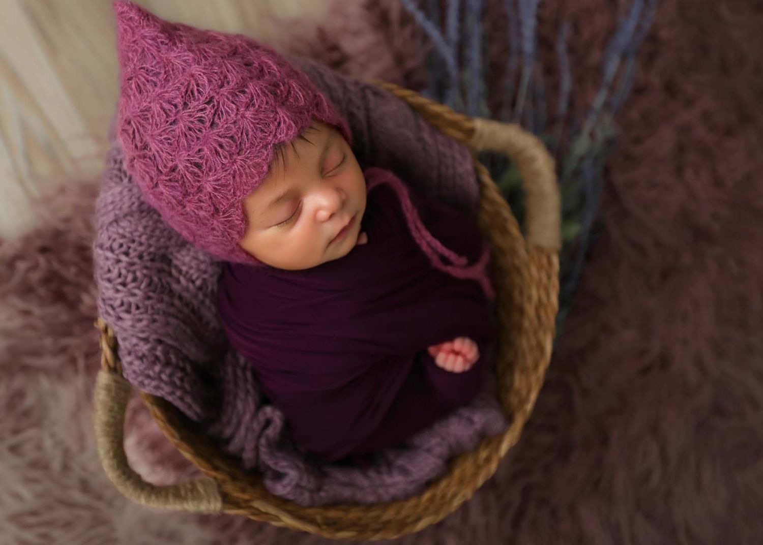 Newborn baby wrapped in purple swaddle wearing pink knit hat sleeping in wicker basket on fur blanket.