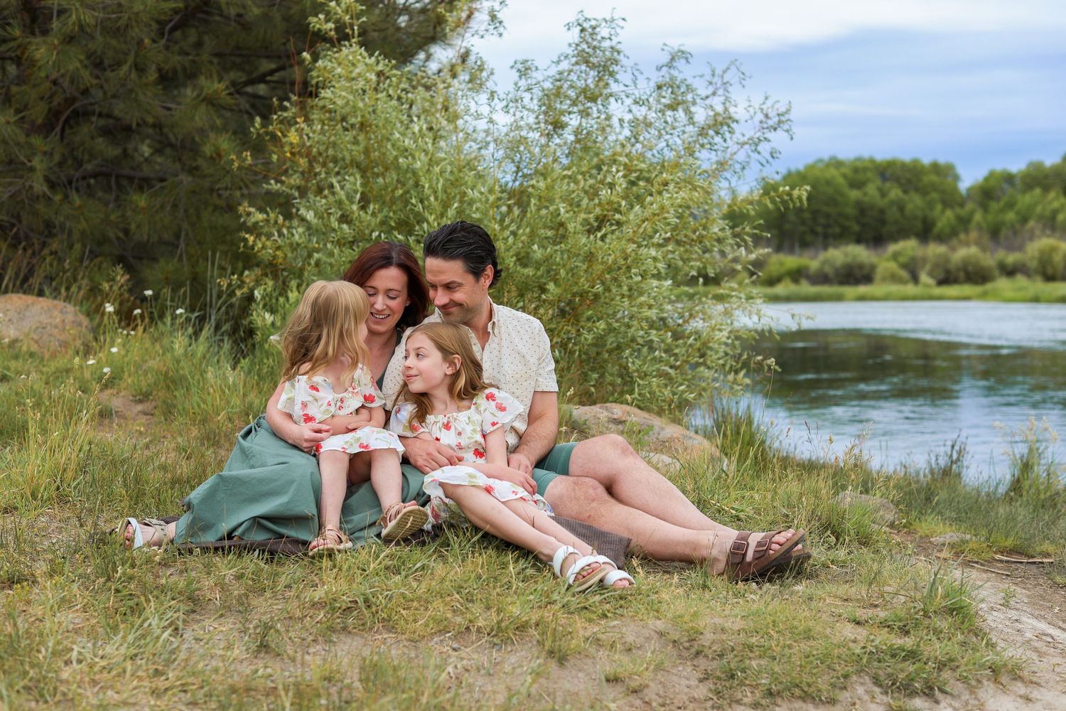 Family of four sitting together on grass by Dillons falls river, surrounded by lush green trees on a sunny day. Bend Oregon