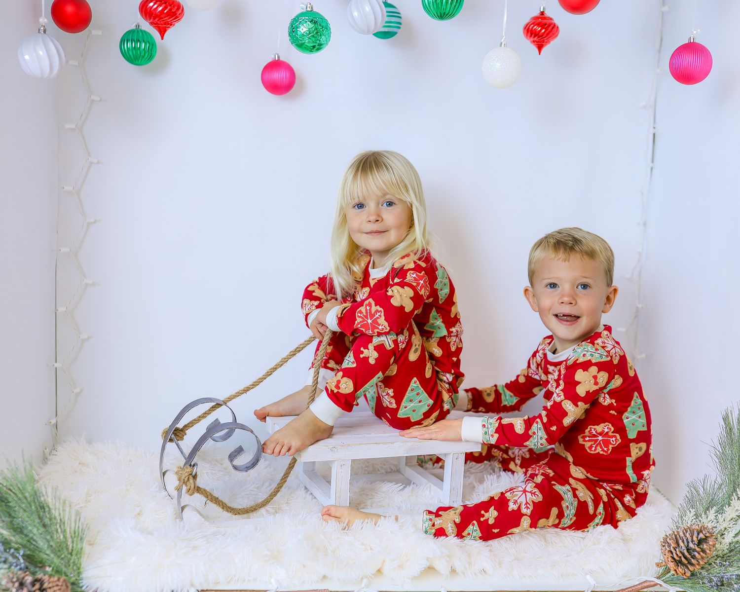 Two smiling children in matching red holiday pajamas sit on a white sled on a fluffy rug beneath hanging Christmas ornaments.