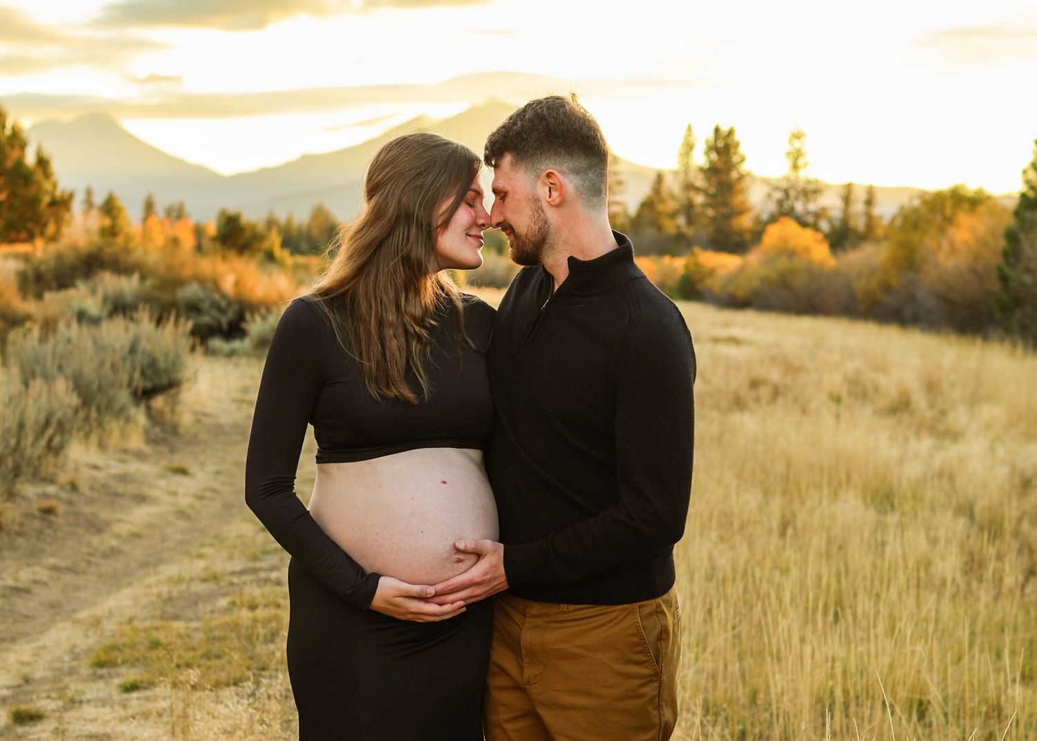 Expecting couple embracing in a golden grassy field at sunset, the woman with her bare baby bump showing, mountains in the background.