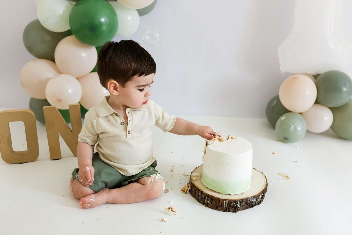 A first birthday cake smash setup with sage green and cream balloons and rustic wood cake stand.