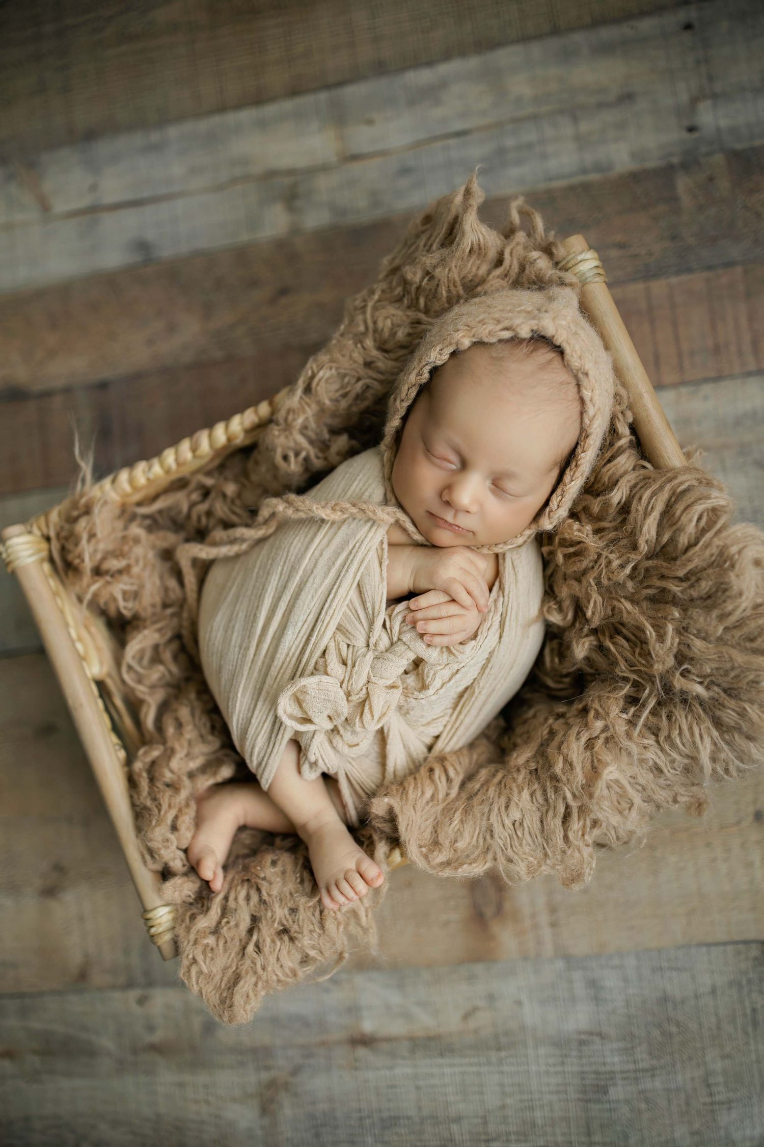 A sleeping newborn in cream-colored wrapping rests on textured fabric against a wooden backdrop.
