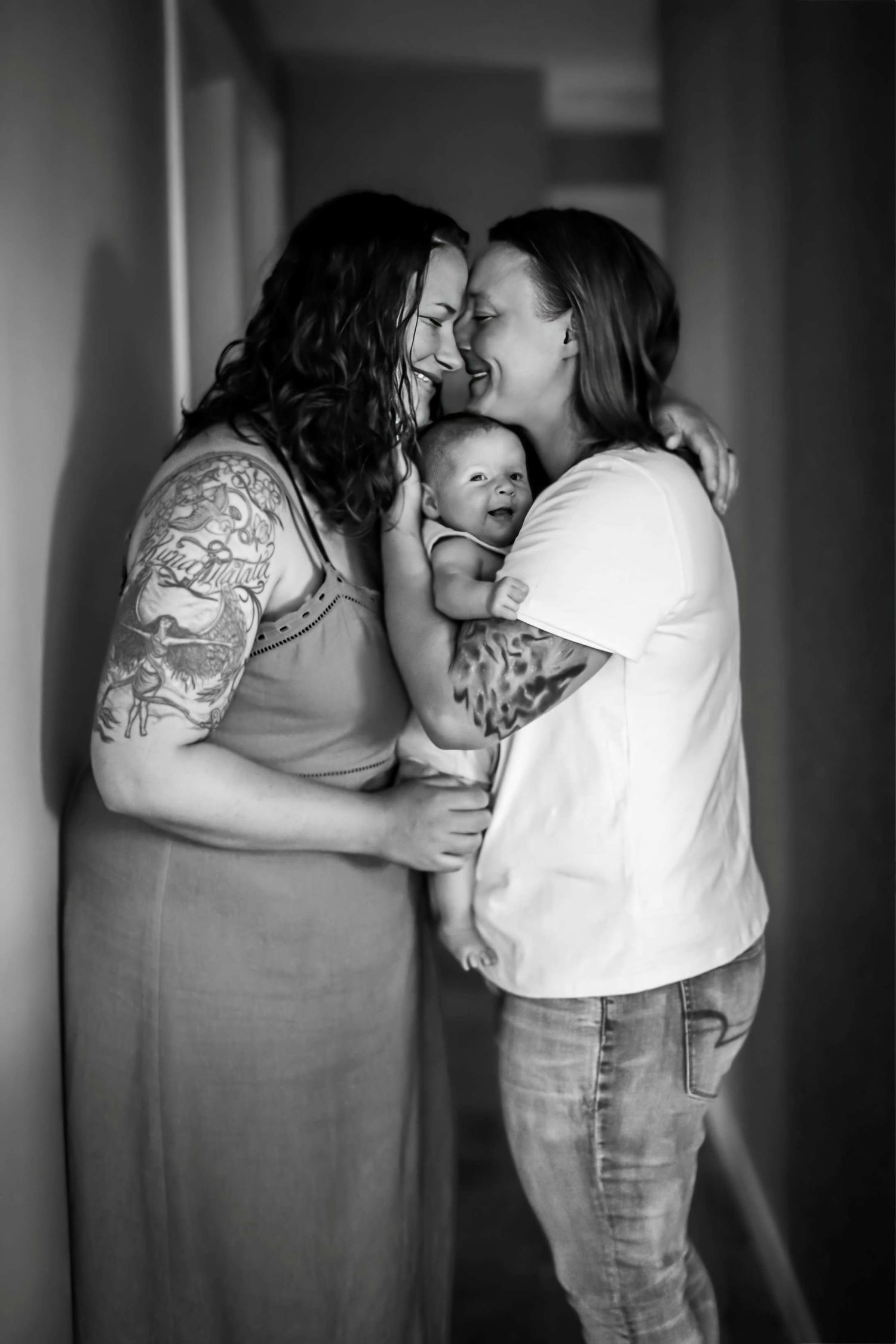Two women share a tender moment smiling at each other while holding their newborn baby in black and white photo.