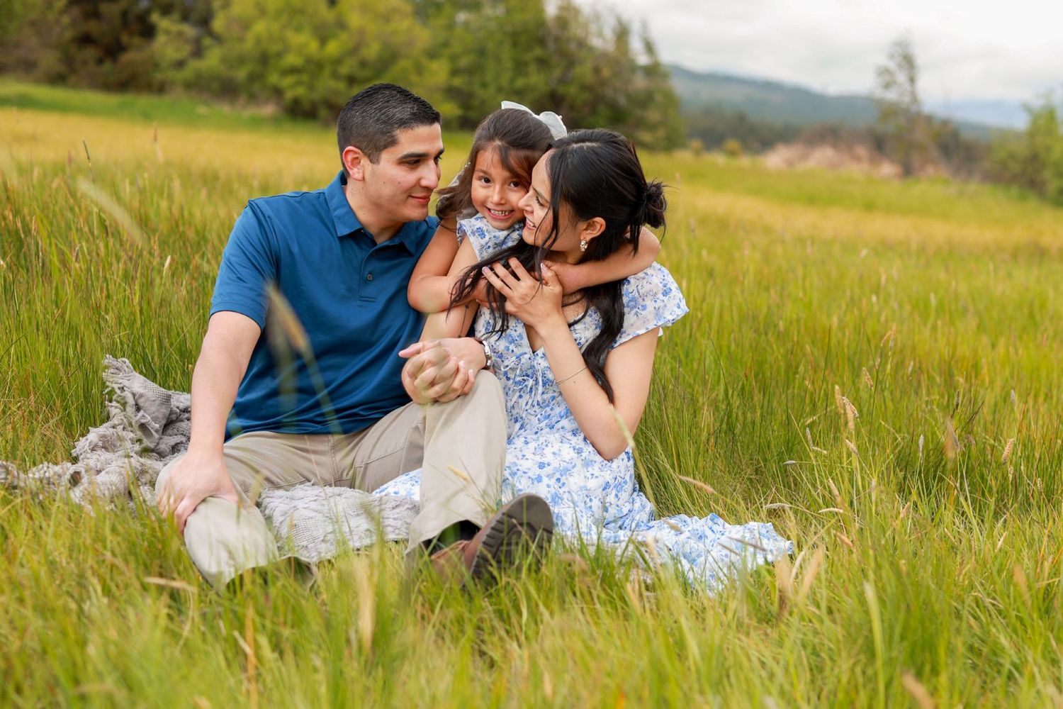 Family of three sitting in a lush green meadow, parents smiling at their young daughter Bend Oregon outdoors.