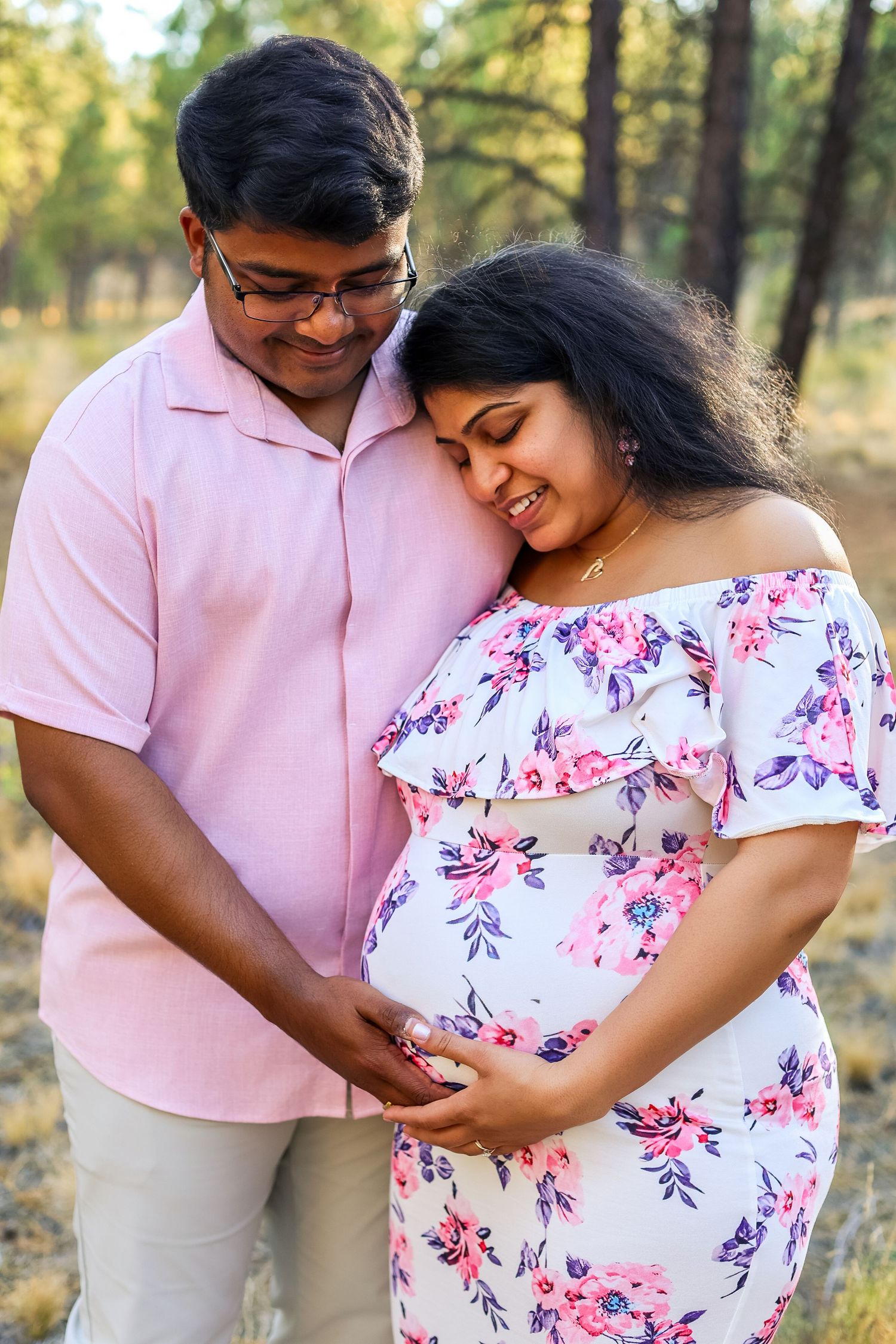 Happy Indian couple in pink floral outfits cradling pregnant belly outdoors in a sunlit forest setting.