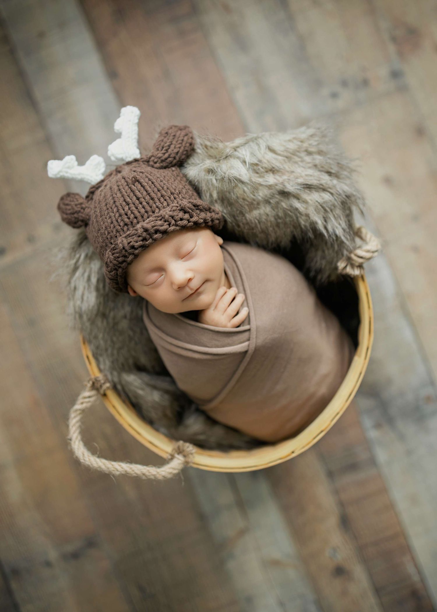 A sleeping baby wearing a brown knit hat lies in a rustic basket with rope handles on wooden flooring.