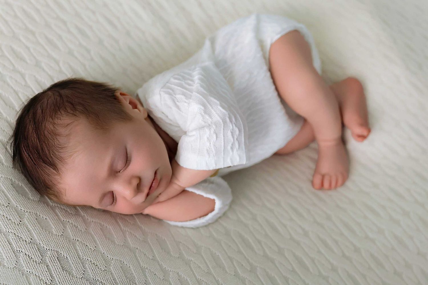 Peaceful newborn baby resting on soft cream-colored blanket in white clothing.