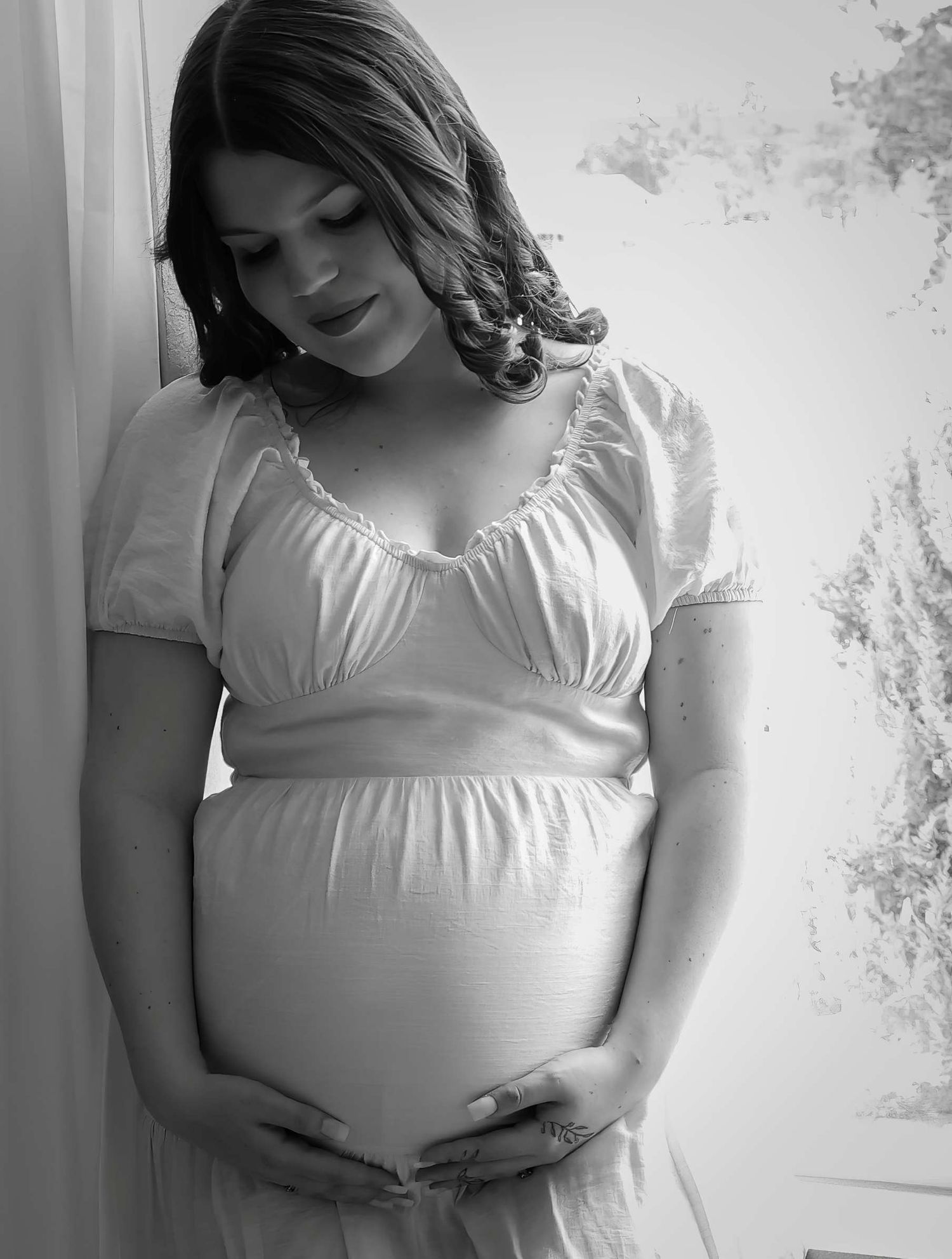 Ethereal maternity portrait in white dress by window with soft natural lighting.