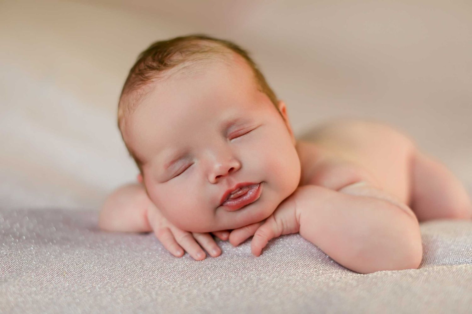 Sleeping baby with rosy cheeks resting head on hands against light background.