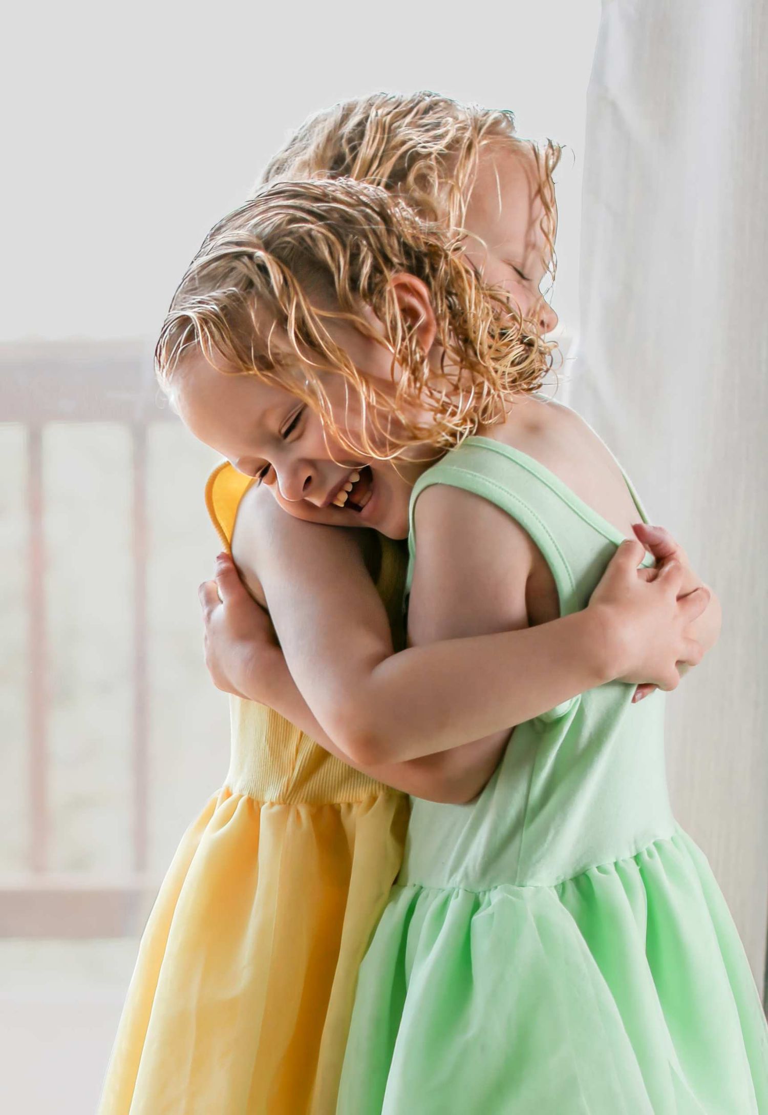 Two children in yellow and mint green dresses share a sweet hug while dancing together in a bright room.
