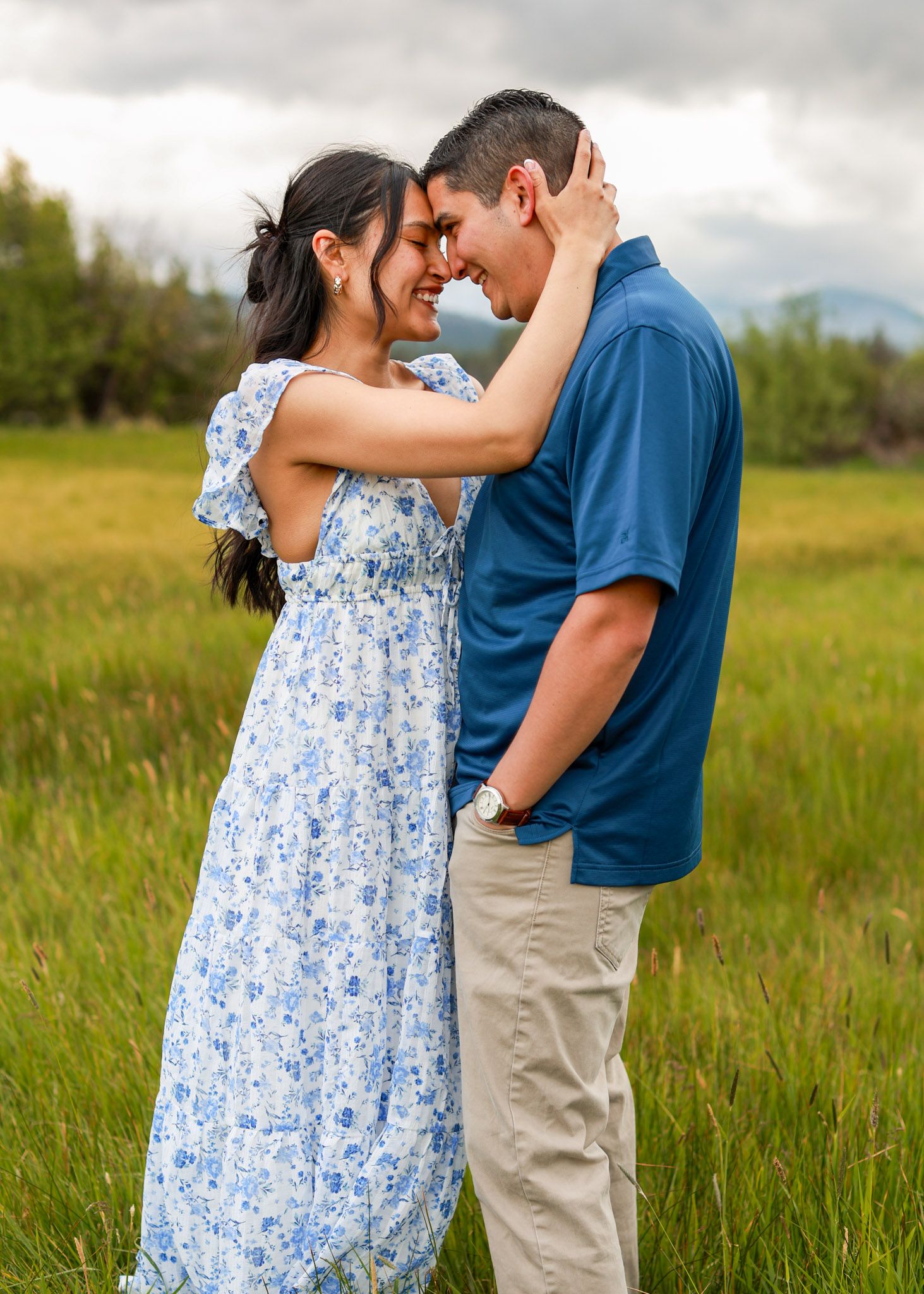 Couple embracing in a green meadow, woman in floral dress touching man's face, mountains in background.