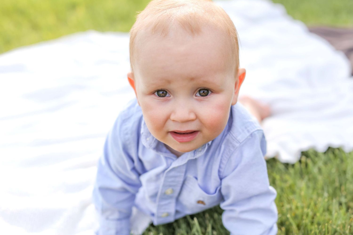 Baby wearing light blue shirt smiles while crawling on white blanket in grass. Black Butte Ranch, Sisters Oregon