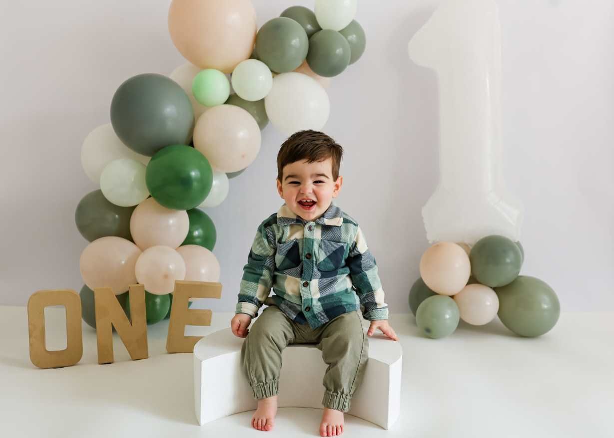 Modern first birthday photo setup with sage green and white balloon arch and gold ONE sign.