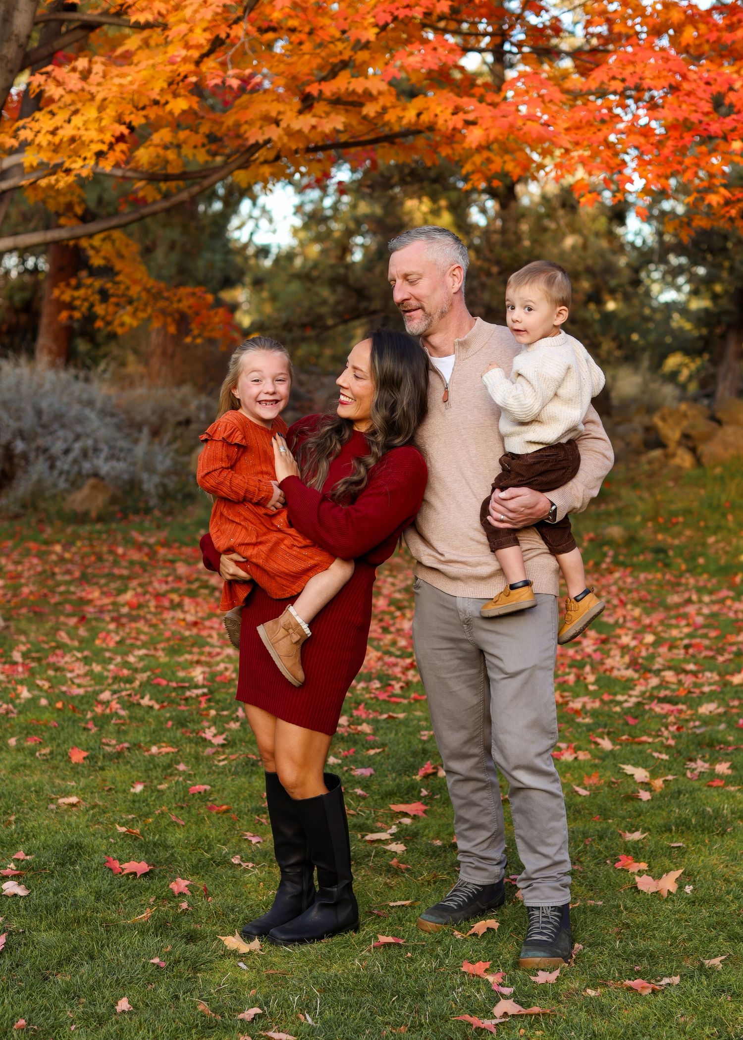 Smiling family of four in coordinated fall outfits stands on a leaf-covered lawn beneath vibrant orange autumn trees.