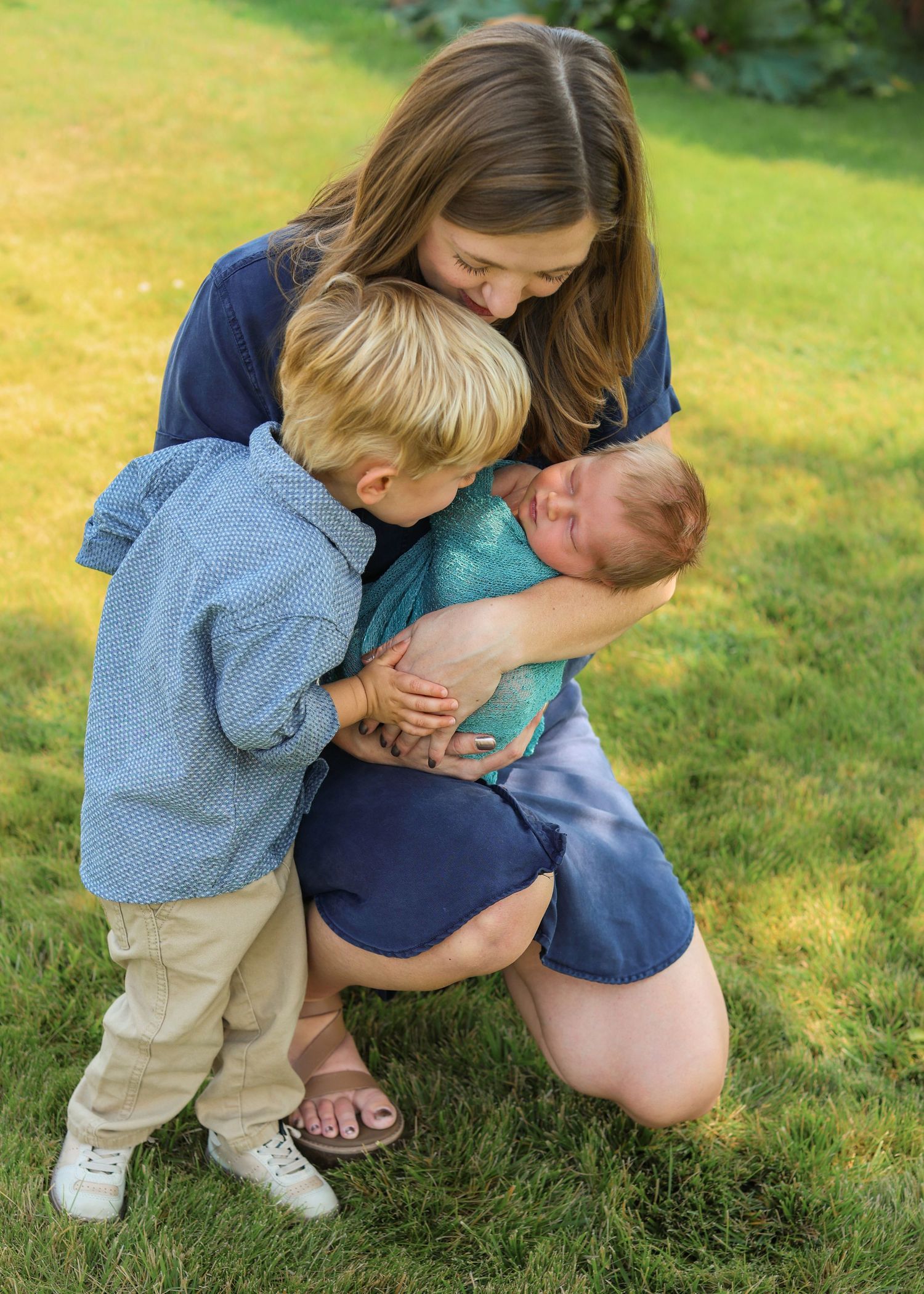A mother kneels on green grass, tenderly holding a newborn while a young toddler boy leans in to look at the baby.
