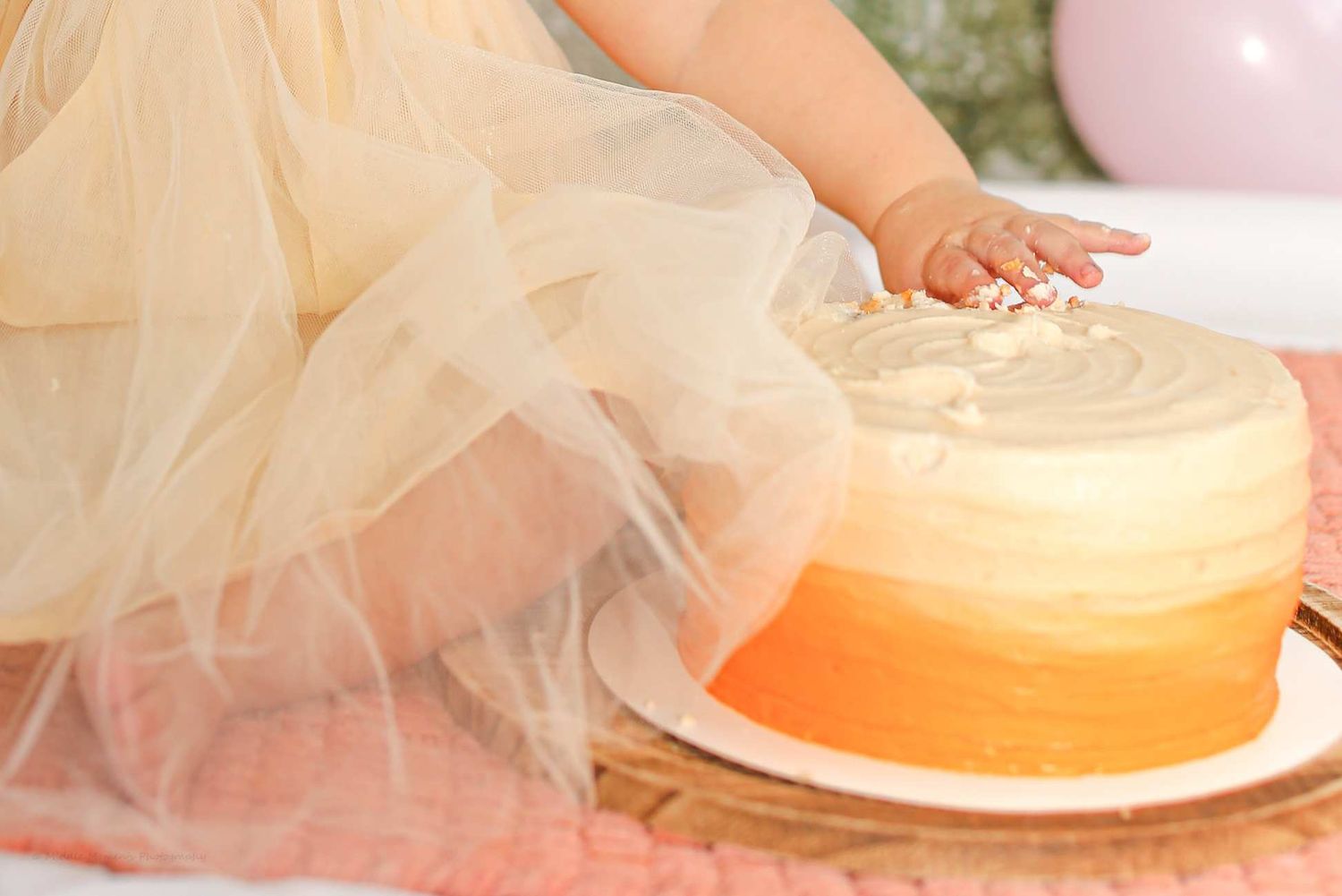 Baby girl in yellow tulle dress smashes orange ombre first birthday cake with her hand.