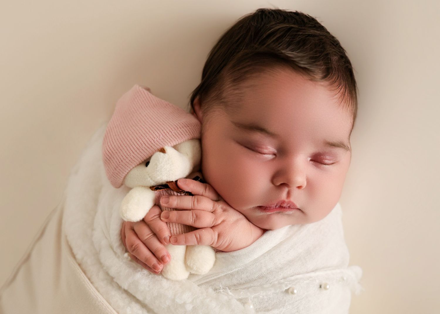 Serene newborn baby swaddled in white lace wrap, sleeping while holding a small white teddy bear with pink hat.