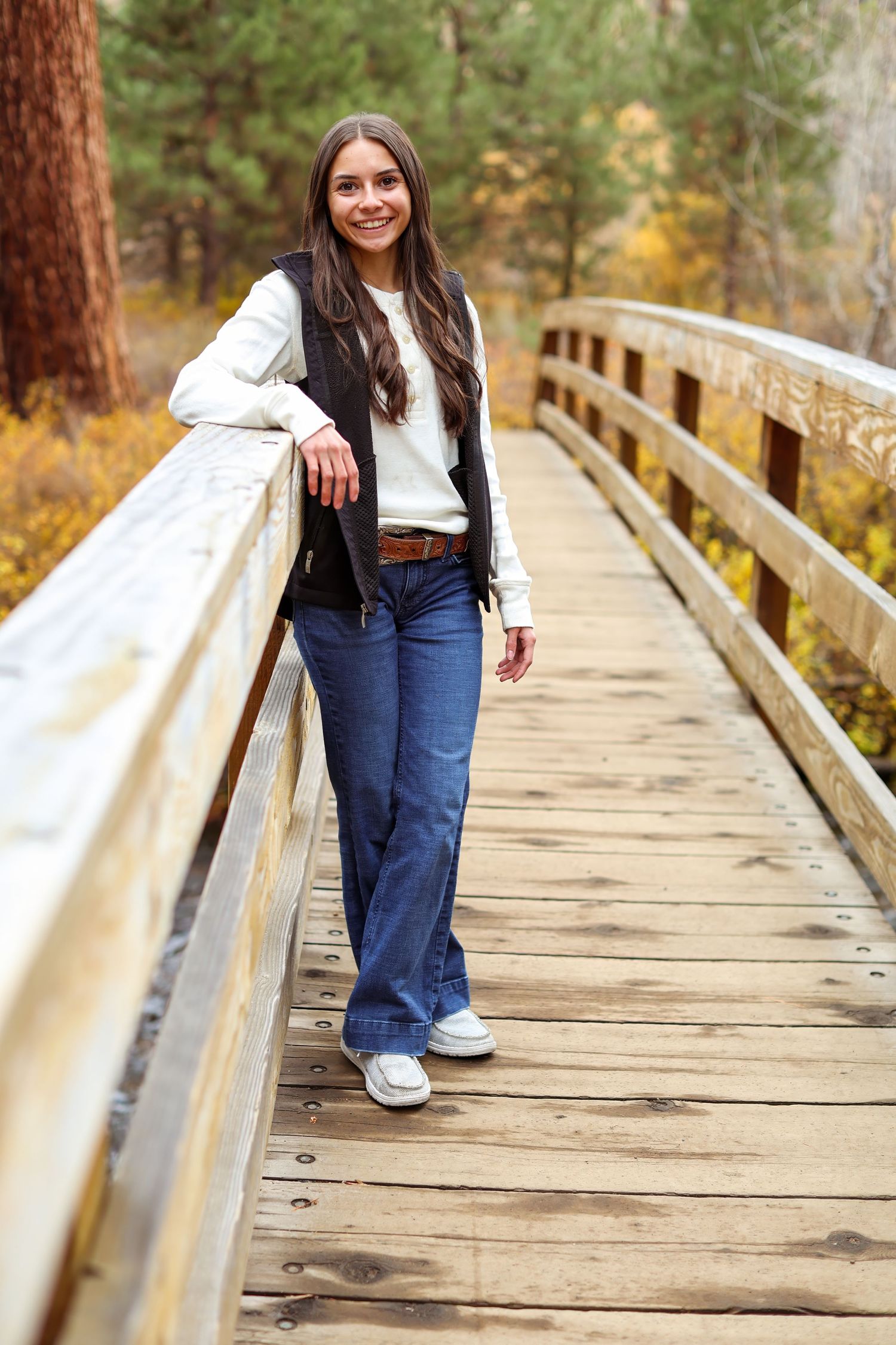 Young woman smiling on a wooden bridge in autumn forest, wearing jeans, white top, and dark vest.