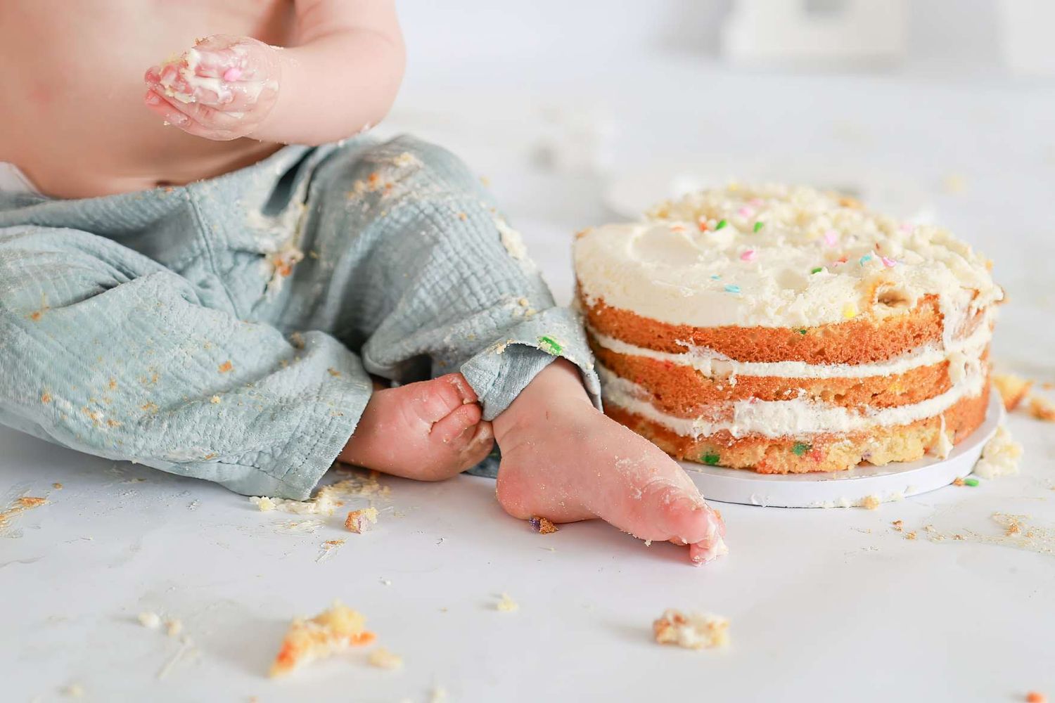 Baby in denim overalls smashing a layered white frosted cake, with crumbs scattered on a white surface. Redmond Oregon Studio