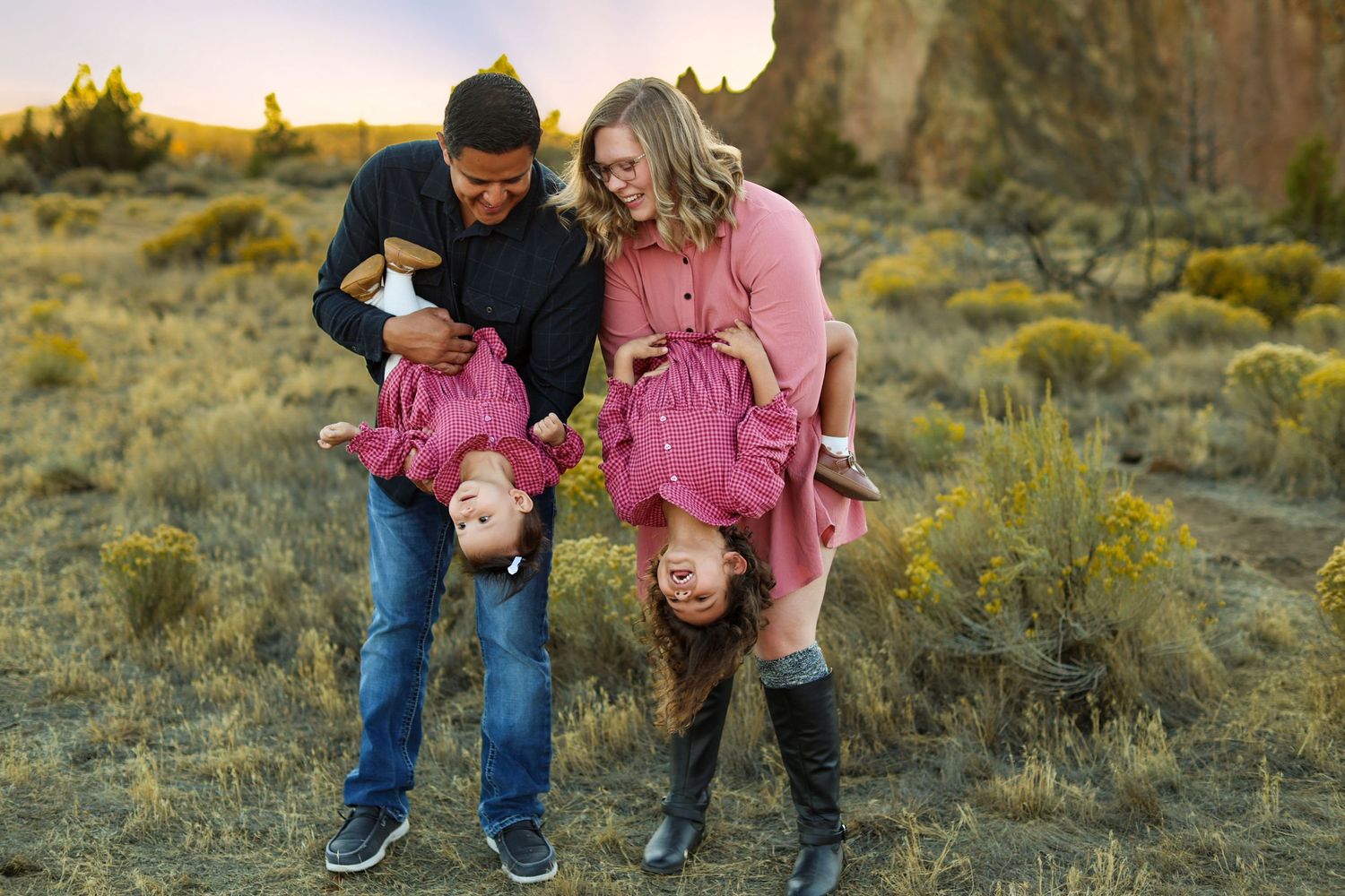 Family of four playfully holding children upside down in a scenic desert landscape at golden hour.