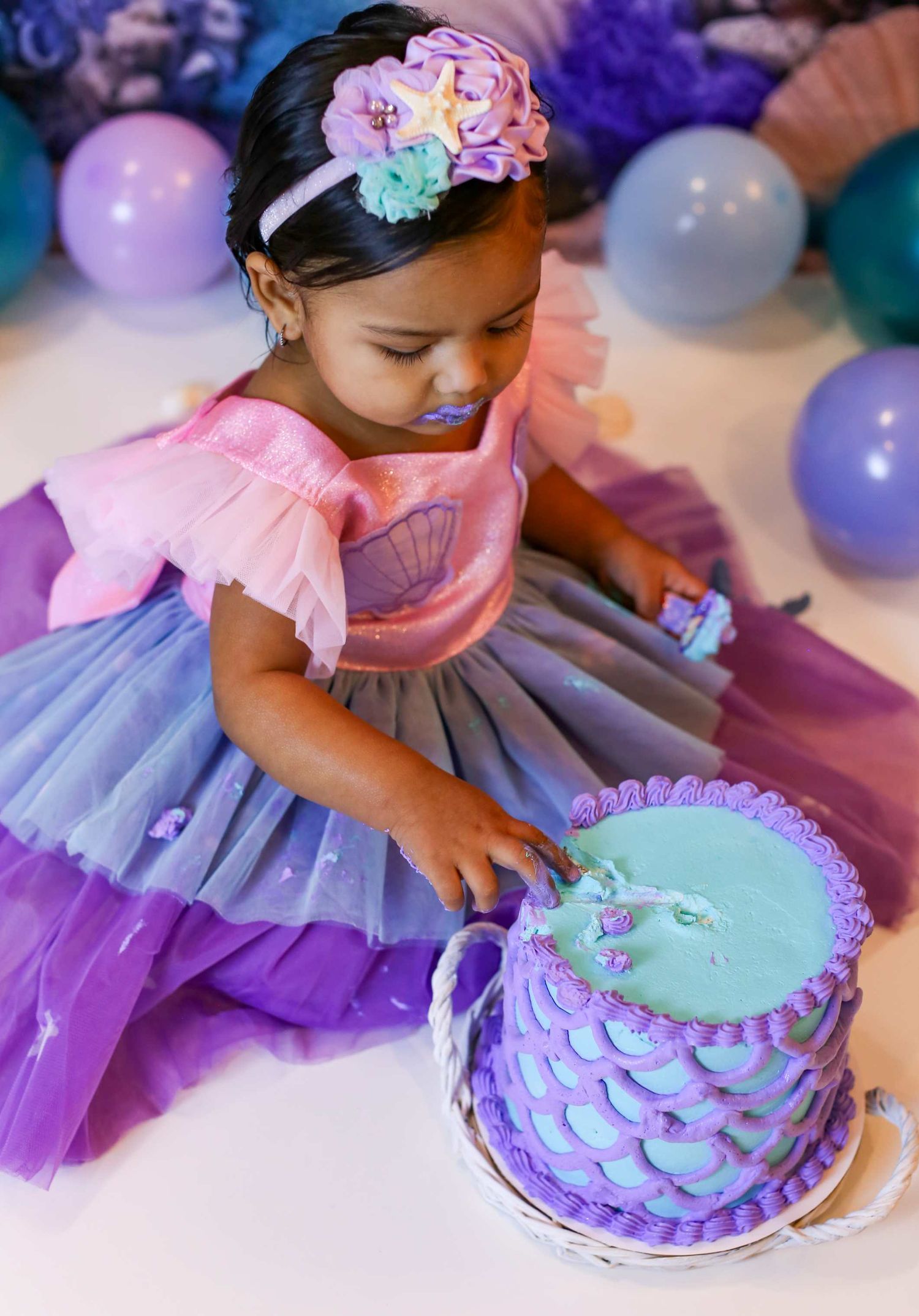 A colorful cake smash photoshoot featuring a pastel purple and teal cake with delicate scalloped details and floral accents.
