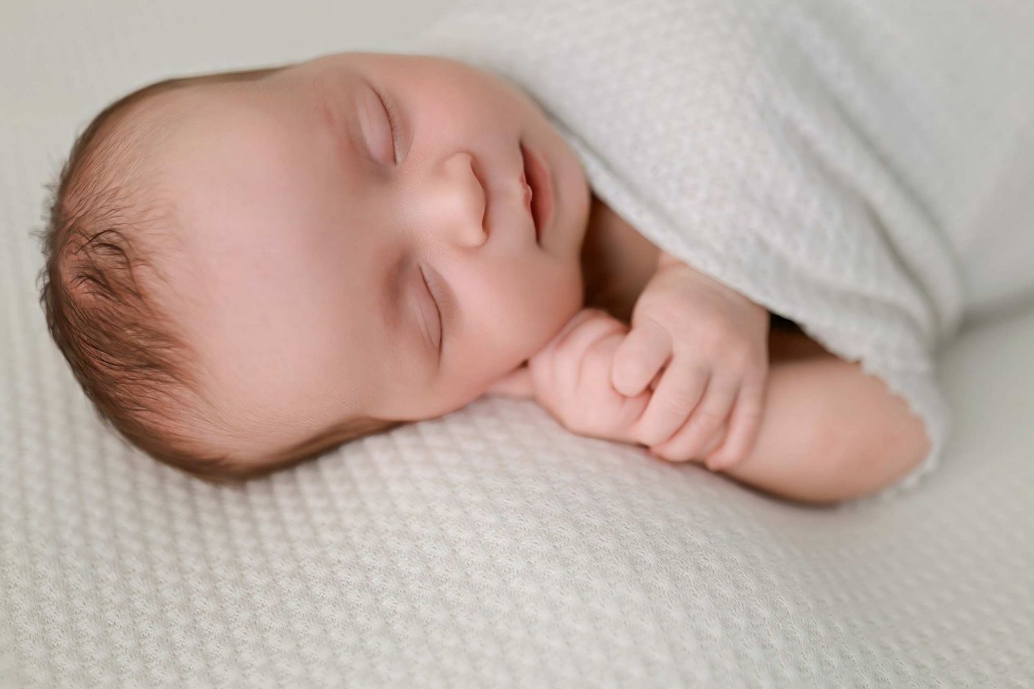 Close-up view of a sleeping newborn baby swaddled in white against a textured background.