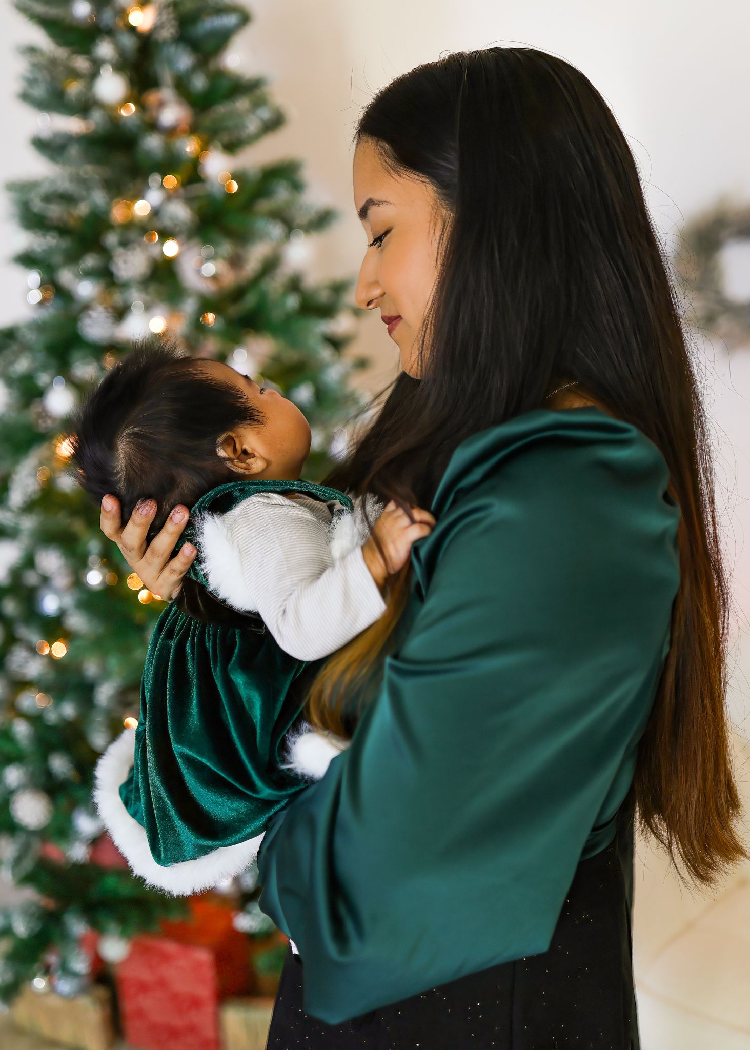 Smiling woman with long dark hair holds a baby in a green velvet dress beside a decorated Christmas tree and wrapped gifts.