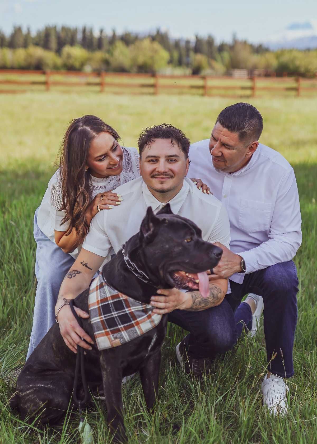 A group in white shirts sits together with their black dog in a grassy field on a sunny day.