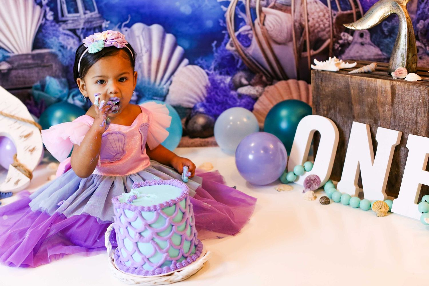 First birthday celebration setup with purple tulle dress, cake, balloons and seashell decorations against blue backdrop.