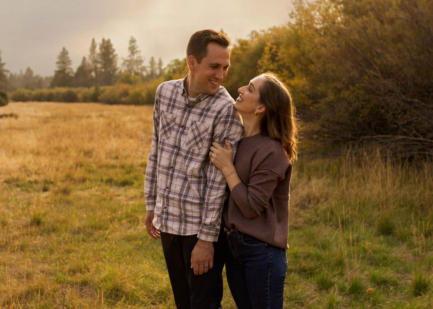 A couple shares tender moments in a golden field during sunset while wearing casual fall attire.