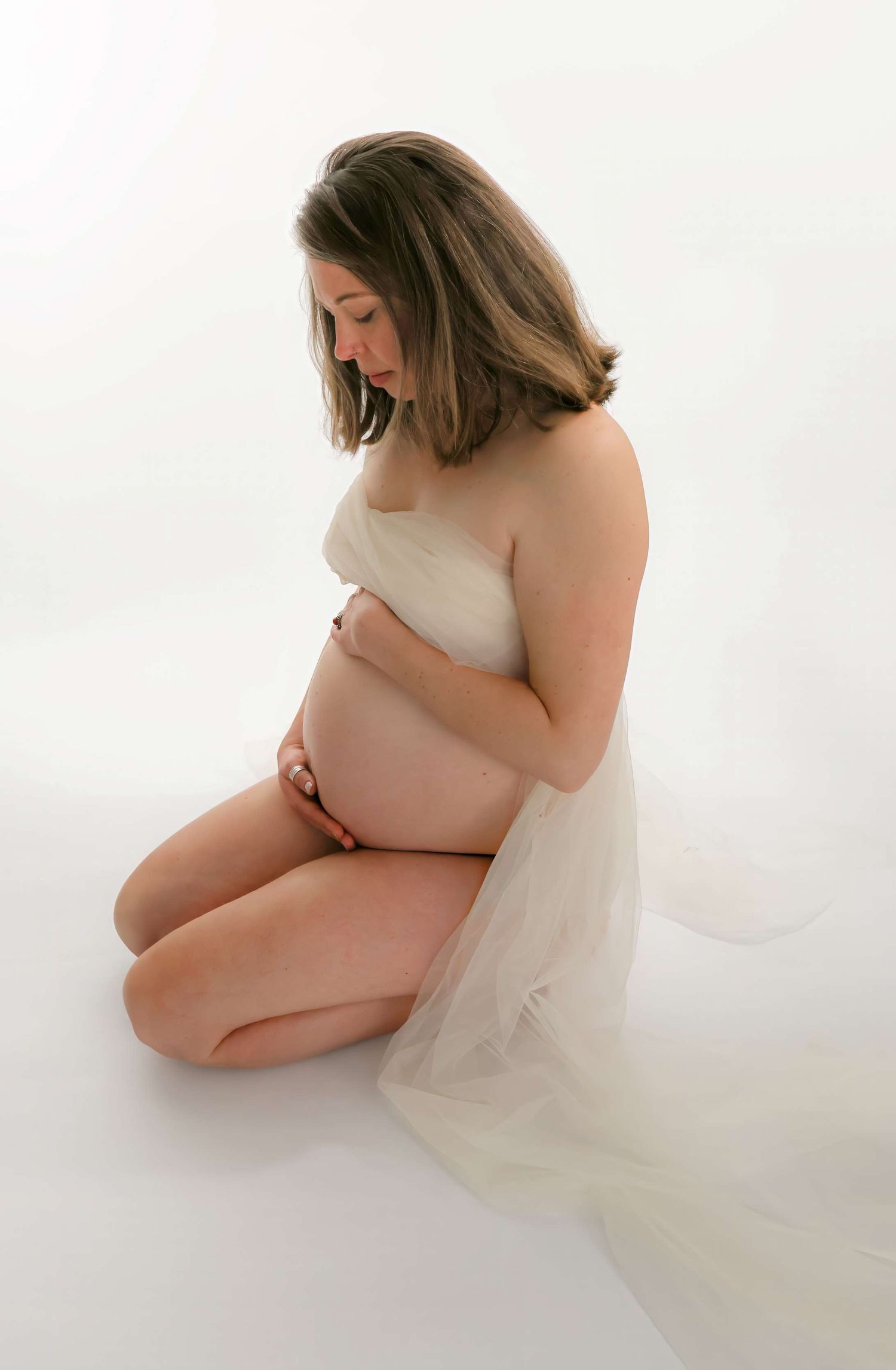 Artistic maternity photo of an expectant mother draped in white fabric against light background.