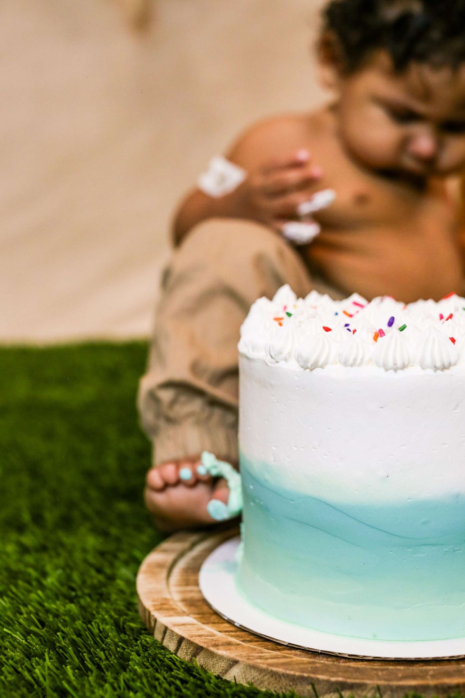 Baby's first birthday smash cake with white and blue frosting on wooden board with infant sitting behind.