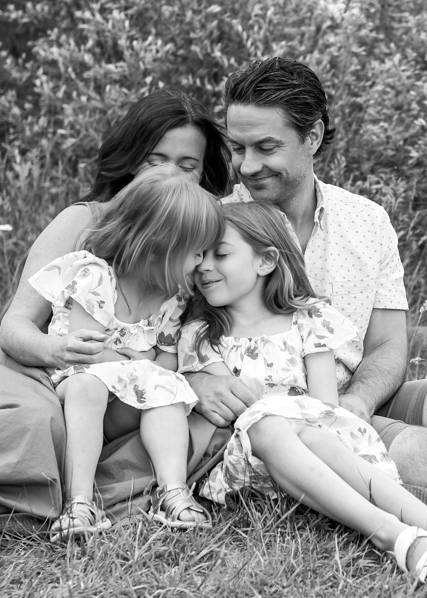 Black and white family portrait of parents and two young daughters sitting together in a field, sharing a tender moment.