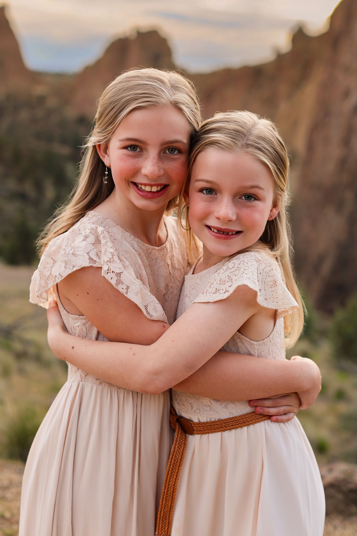 Two smiling blonde girls in lace dresses embrace warmly with rocky desert cliffs in the background. Smith Rock State Park