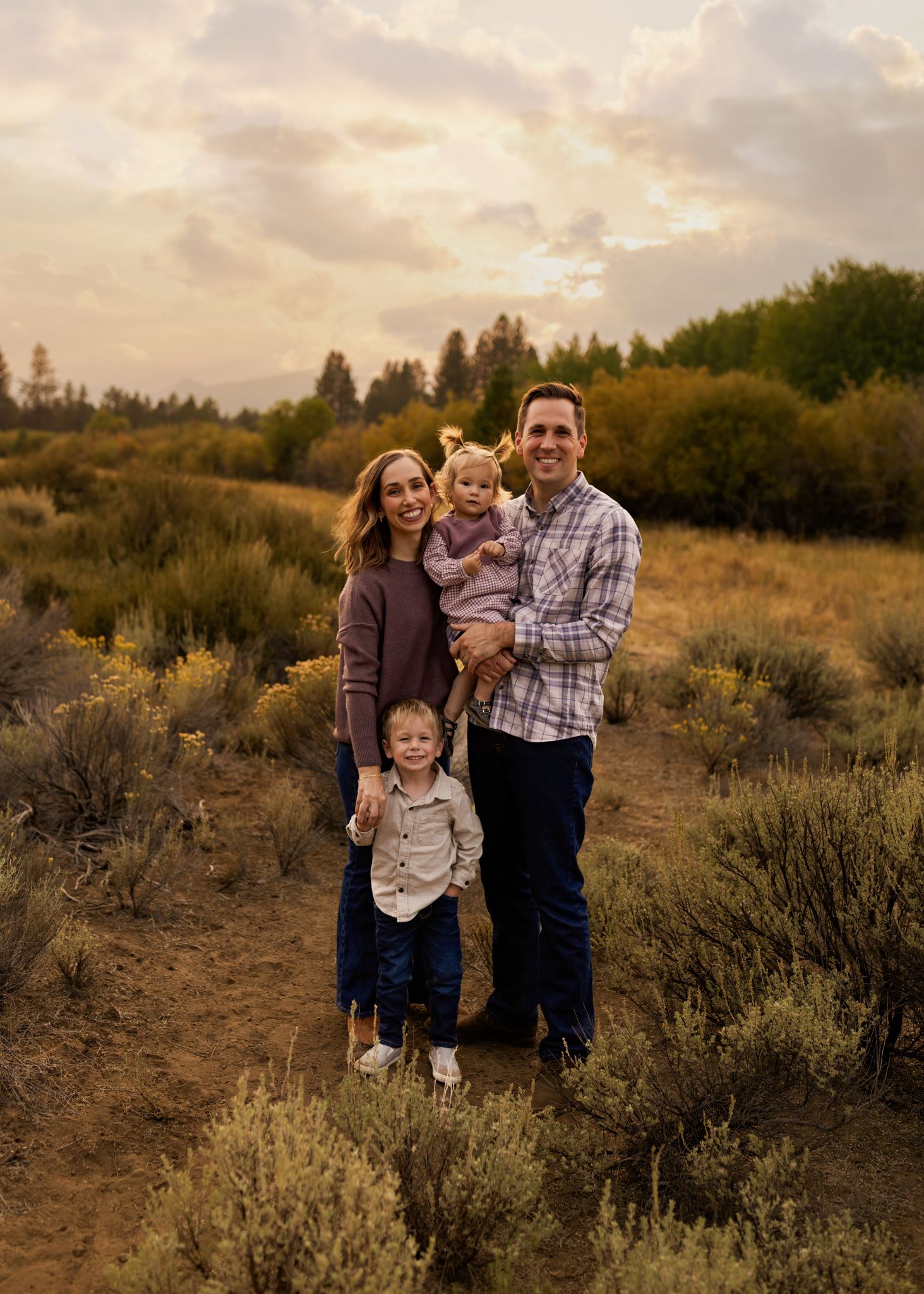 Happy family of four poses at golden hour in scenic sagebrush field with cloudy sky.