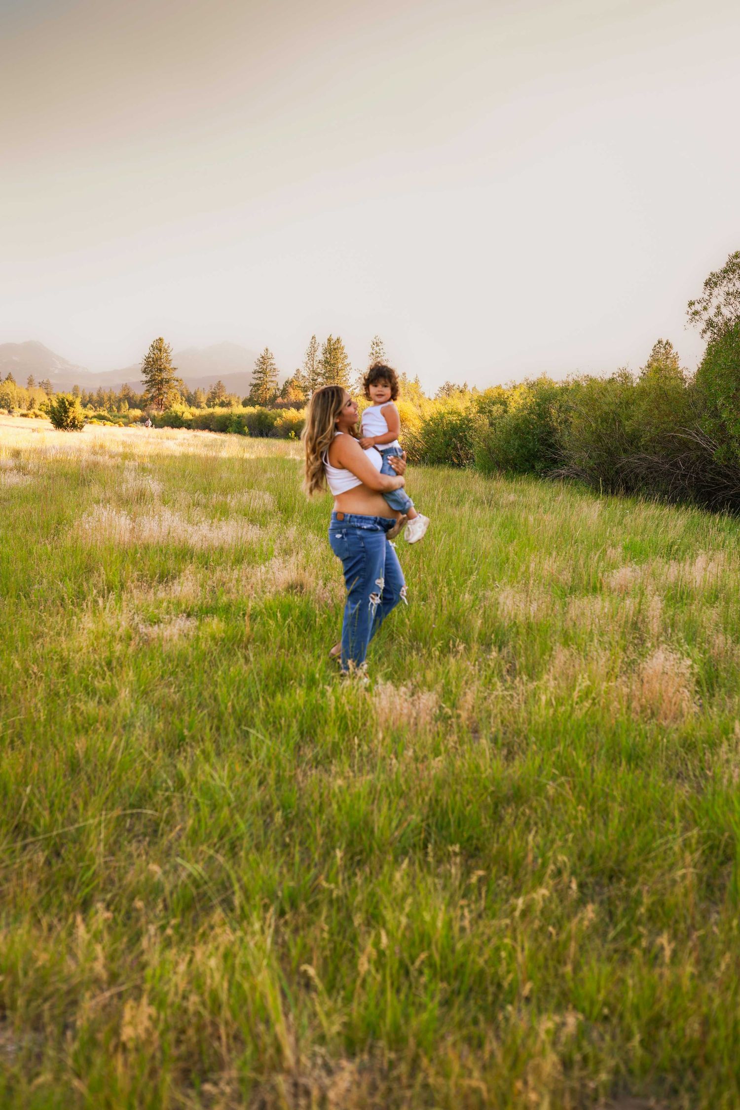 A mother in jeans and white top holds her young child in a golden sunlit meadow.